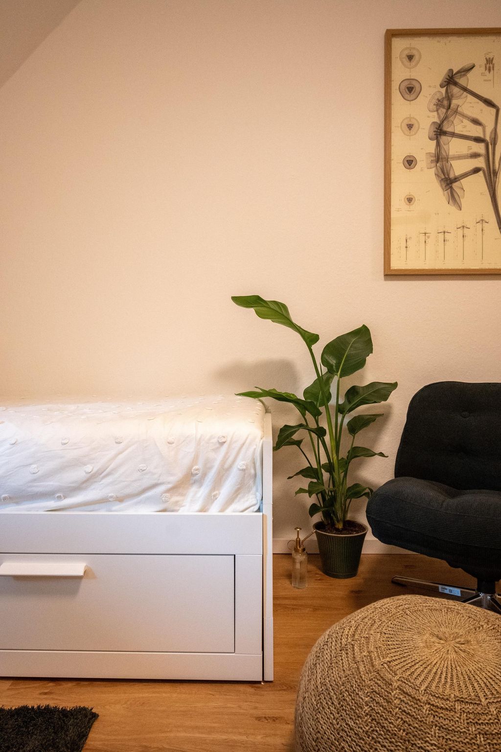 A minimalist bedroom with a white bed featuring cotton bedding, a green potted plant next to a black office chair, and a linen pouf on a wooden floor against a cream-colored wall.