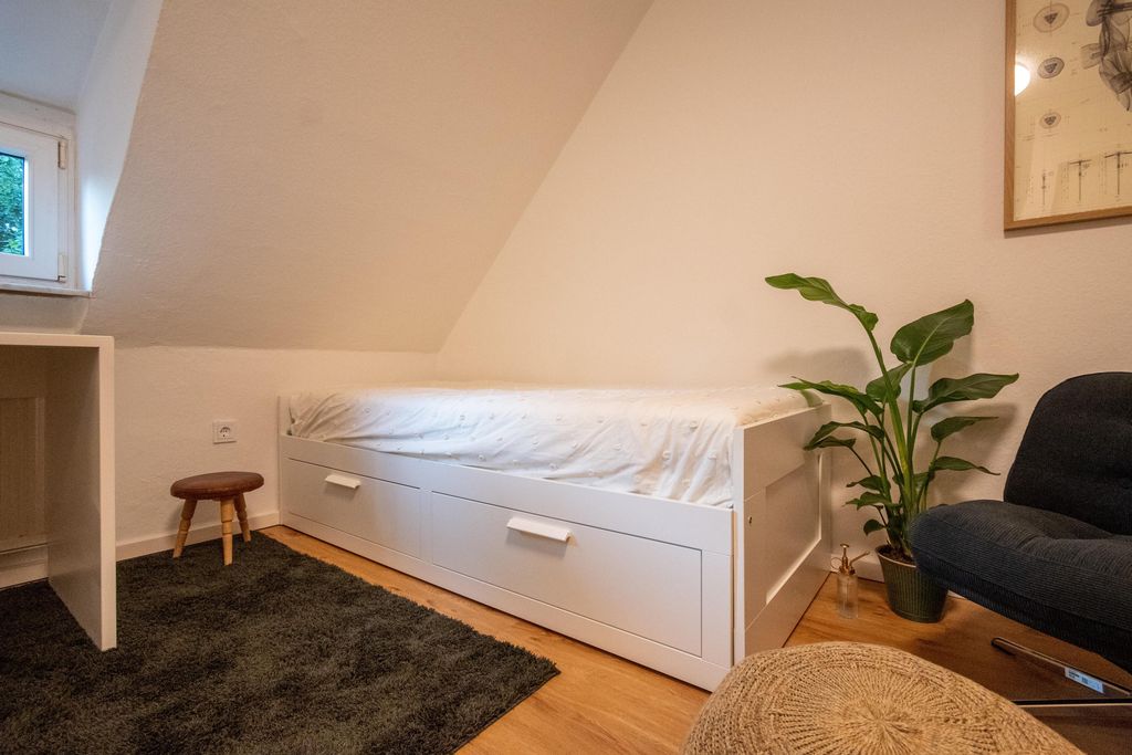 A minimalist bedroom under a sloped ceiling with a white bed, black rug, small stool, potted plant, and dark armchair.