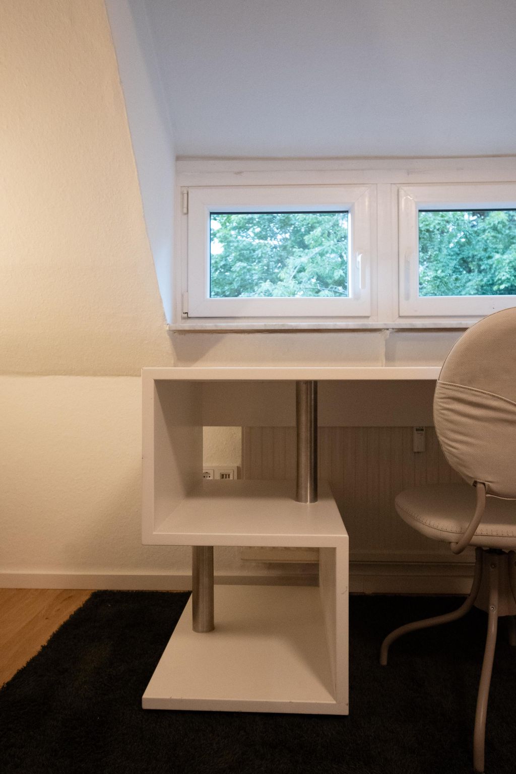 White, modern geometric side table shelf made of wood with metal supports in front of a small window showing green trees outside.
