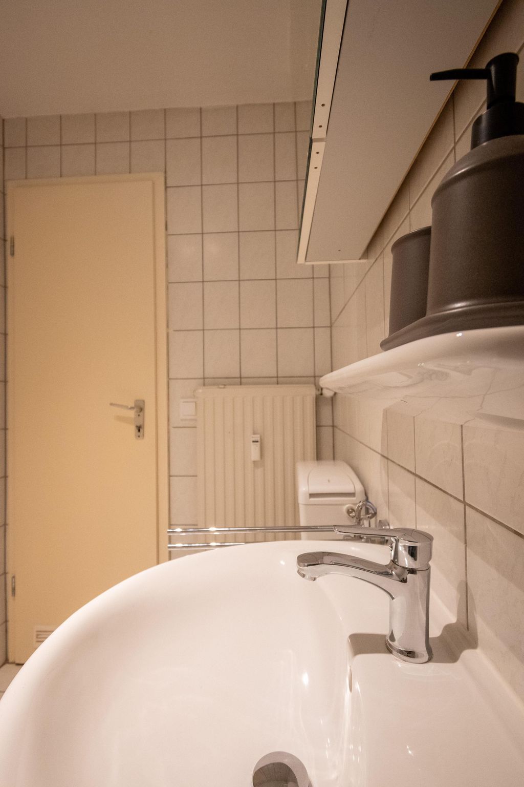 A bathroom with a white sink and chrome faucet, square tiled wall, beige door, radiator, and soap dispenser on the right side.