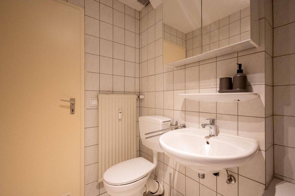 Small bathroom with white toilet and sink, beige tiled walls. A mirror with a shelf holding two soap dispensers is mounted on the wall.