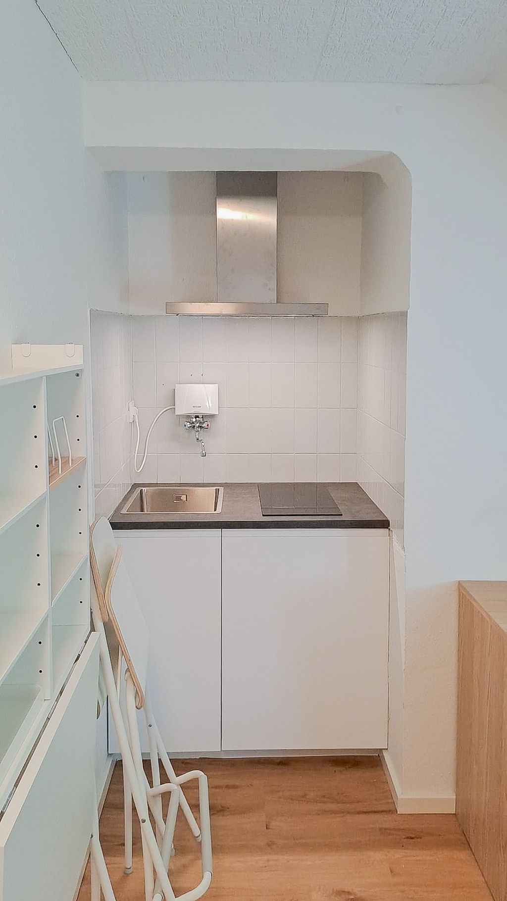 Small kitchenette with white tiled backsplash, sink, stovetop, and stainless steel range hood in a recessed area, with two folded chairs in front.