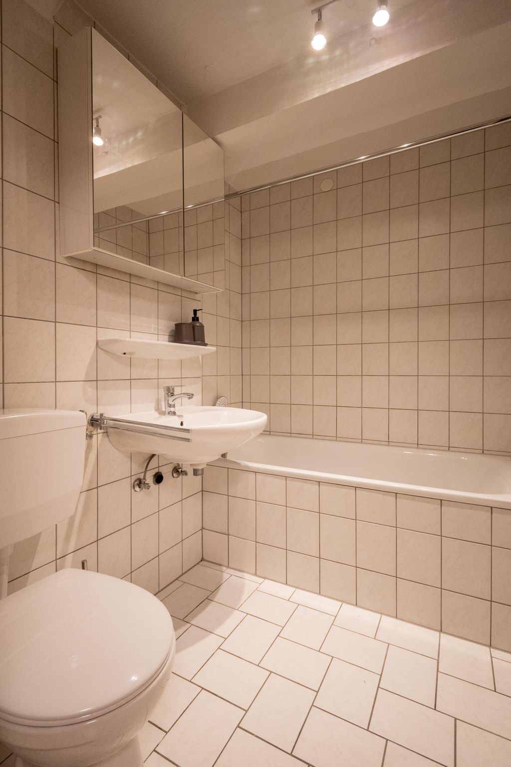 Small bathroom with white tiles, toilet, wall-mounted sink with faucet, rounded mirror cabinet, soap dispenser, and bathtub.