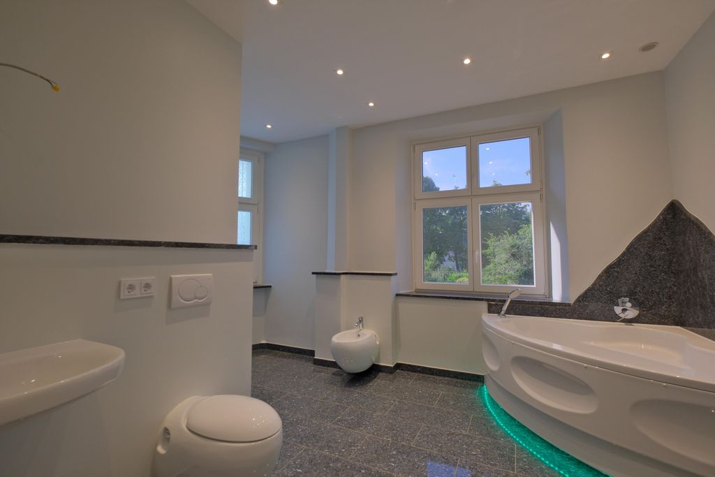 A modern bathroom with gray tiled floor, white walls, a corner bathtub with green LED lighting, sink, toilet, and bidet, in front of a large window overlooking greenery.