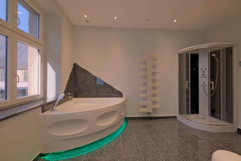 A modern bathroom featuring a curved bathtub with green LED lighting underneath, a shower with glass doors, and a white radiator mounted on the wall.