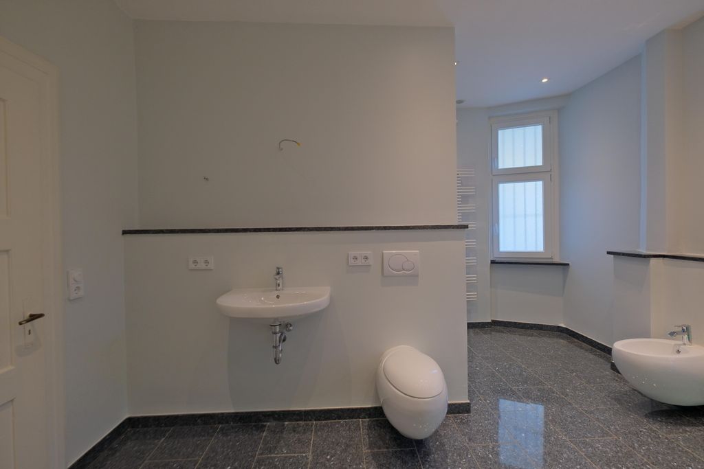 A modern bathroom with white walls, a wall-mounted toilet, a wall-mounted sink, and dark tiled floor. A window and radiator are visible in the background.