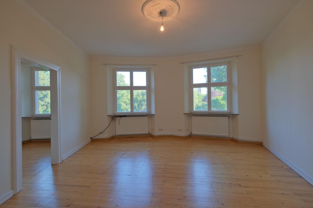 Empty room with light wooden floors, two large windows overlooking trees, and an adjoining doorway to another room with windows.