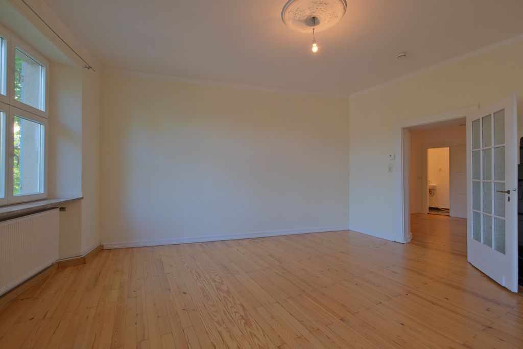 Empty room with wooden floor, white walls, a window on the left with a radiator below, and an open door leading to another room.