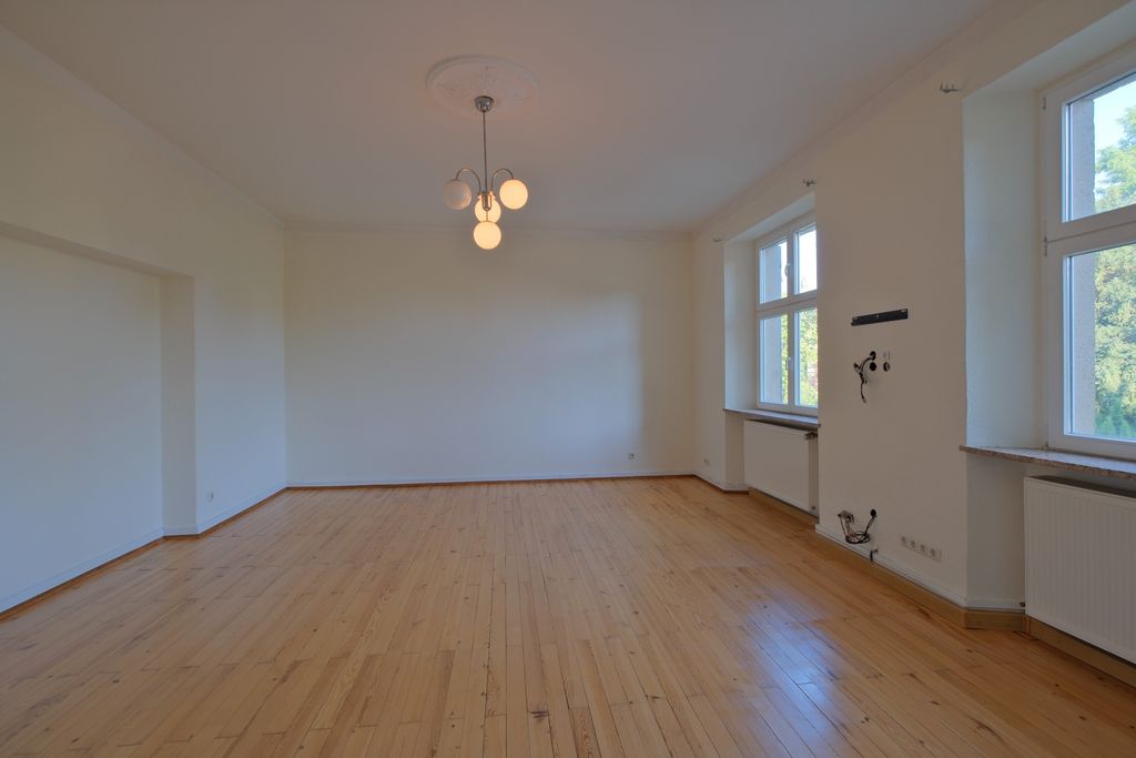 Empty bright room with wooden flooring, large windows on the right side, and a modern ceiling light fixture.