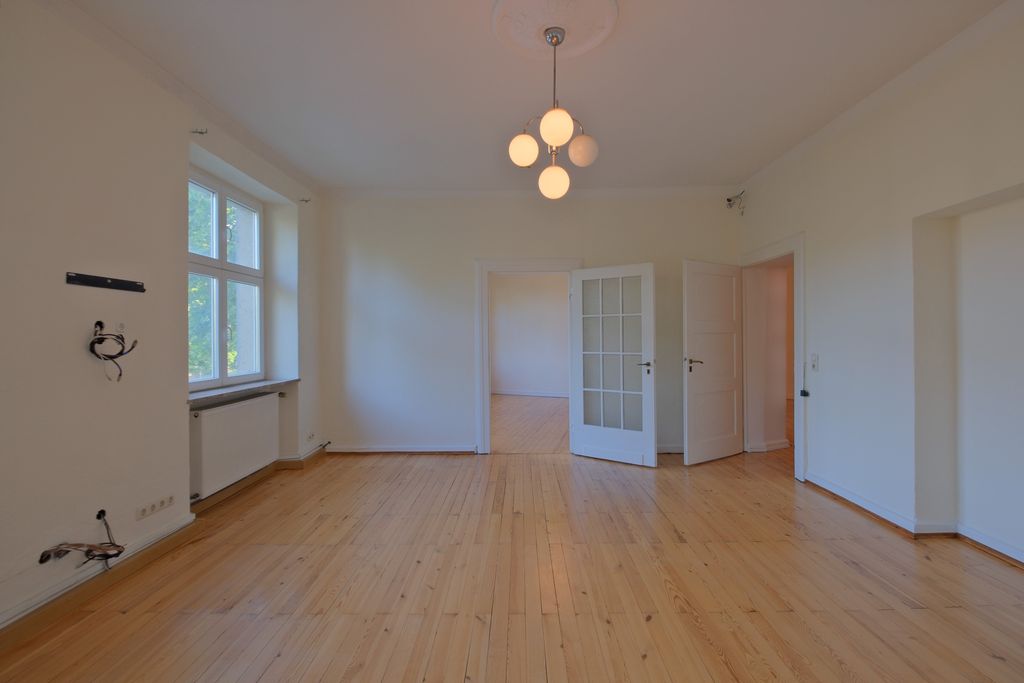 Empty room with light wooden floor, white walls, and a modern ceiling light featuring five round bulbs, with visible doors and window looking outside.