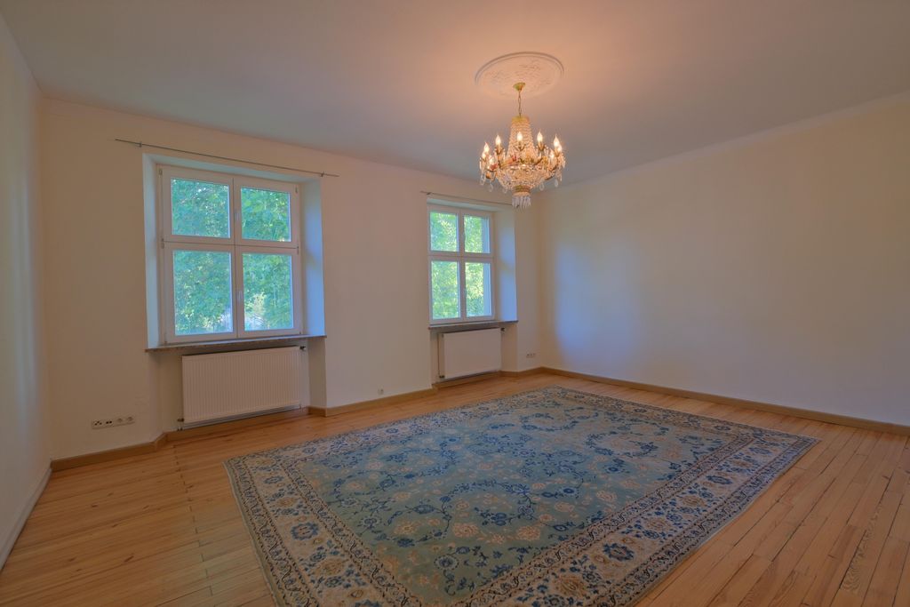 Empty room with light-colored walls, two windows showing daylight and green trees outside, a large decorative carpet on the wooden floor, and a chandelier hanging from the ceiling.