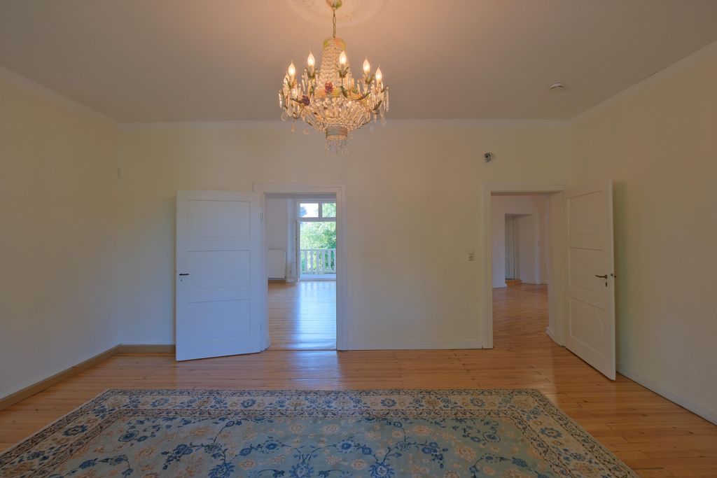Empty spacious room with wooden flooring, a floral carpet, and a chandelier on the ceiling, featuring white doors leading to adjacent rooms.