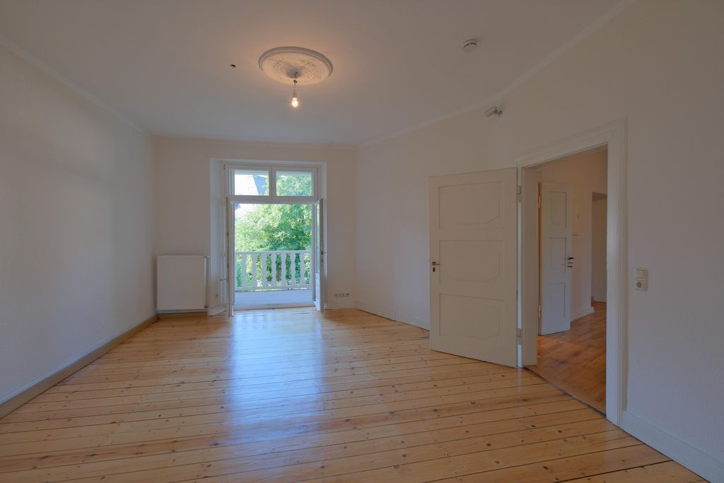 Empty room with wooden floor, open balcony door showing green trees outside, and white walls.