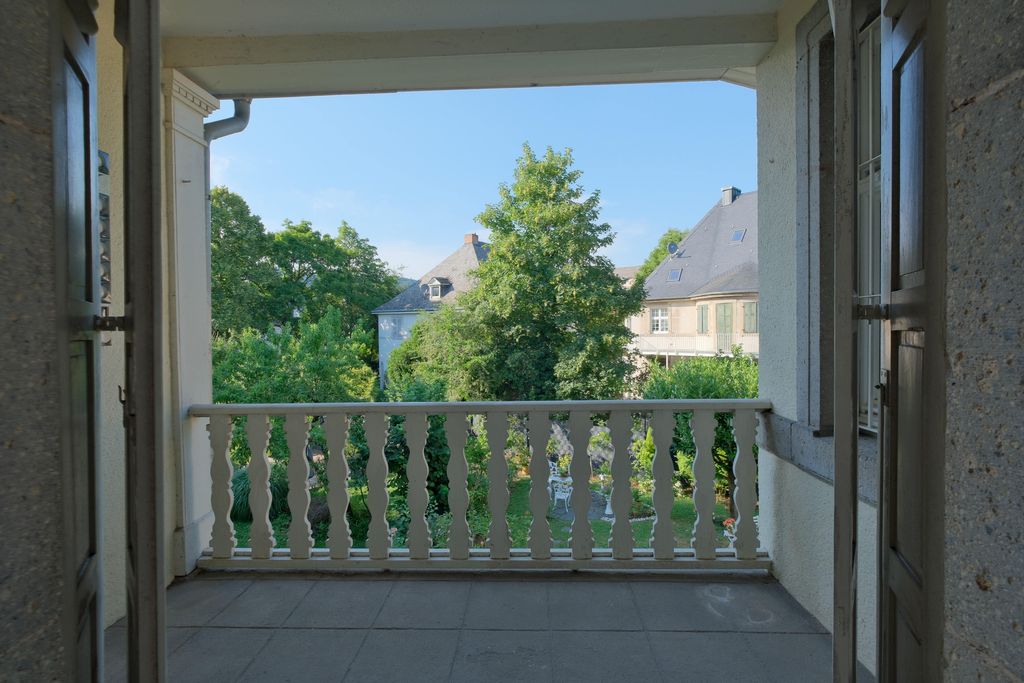 View through an open doorway onto a balcony with a white railing, overlooking a garden with trees and houses in the background under a clear sky.