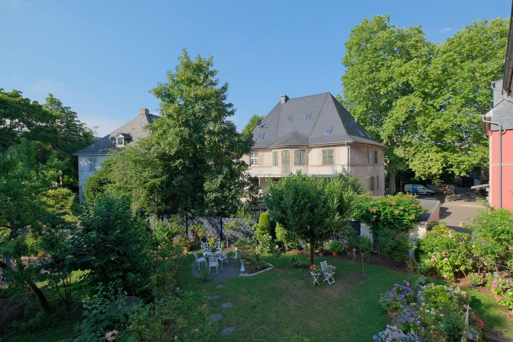 View of a well-kept garden with winding paths, white garden furniture, and blooming plants in front of a large historic house with a steep dark roof and green shutters under a blue sky.