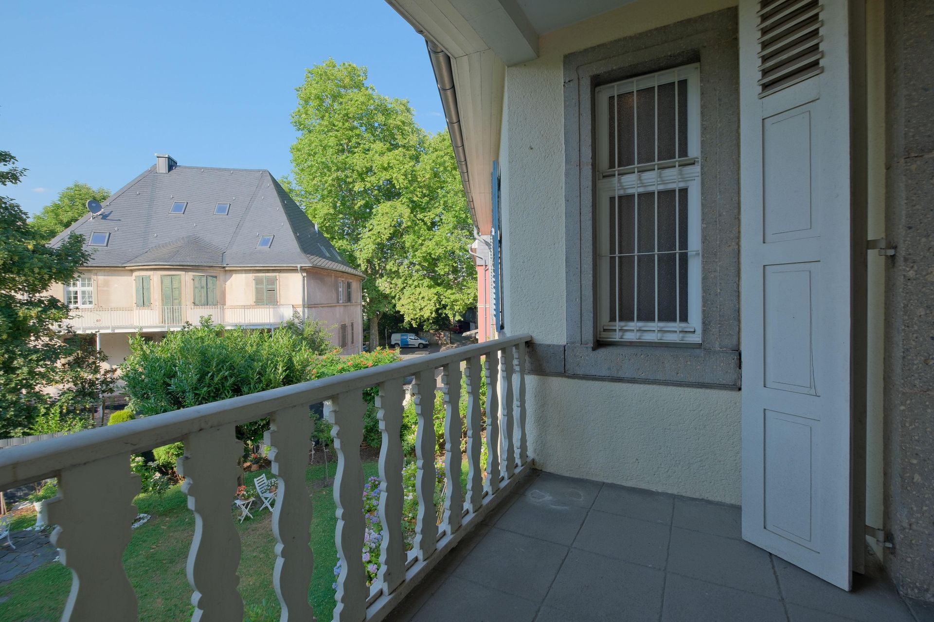 Balkon mit weißem Geländer und Fliesenboden, ein Fenster mit Gitter und eine offene weiße Tür, im Hintergrund ein großes Haus und grüne Bäume unter klarem Himmel.