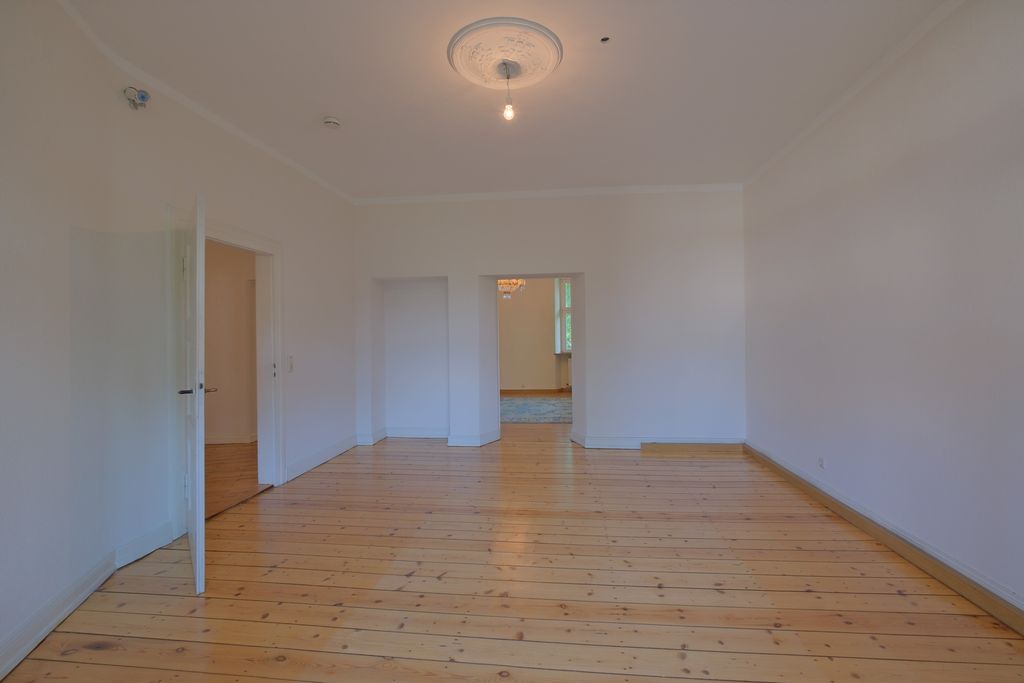 Empty room with white walls and wooden floor, featuring a single ceiling light and open doors leading to another room.