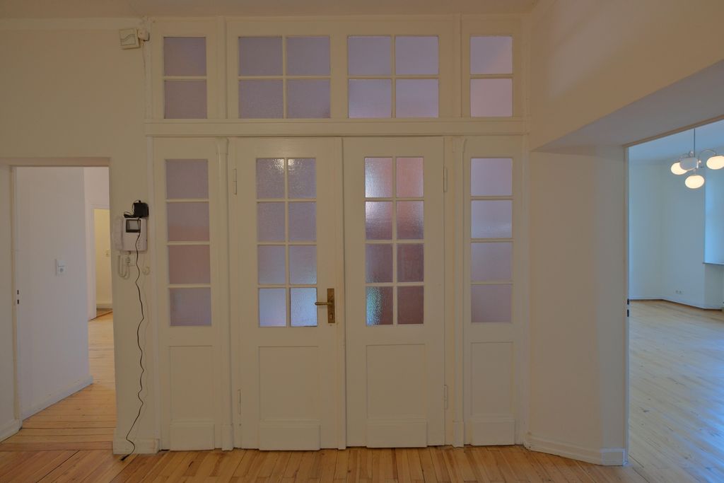 View of a white double door with glazed window sections in a bright room with wooden flooring. Open doorways to other rooms are visible on the left and right sides of the door.