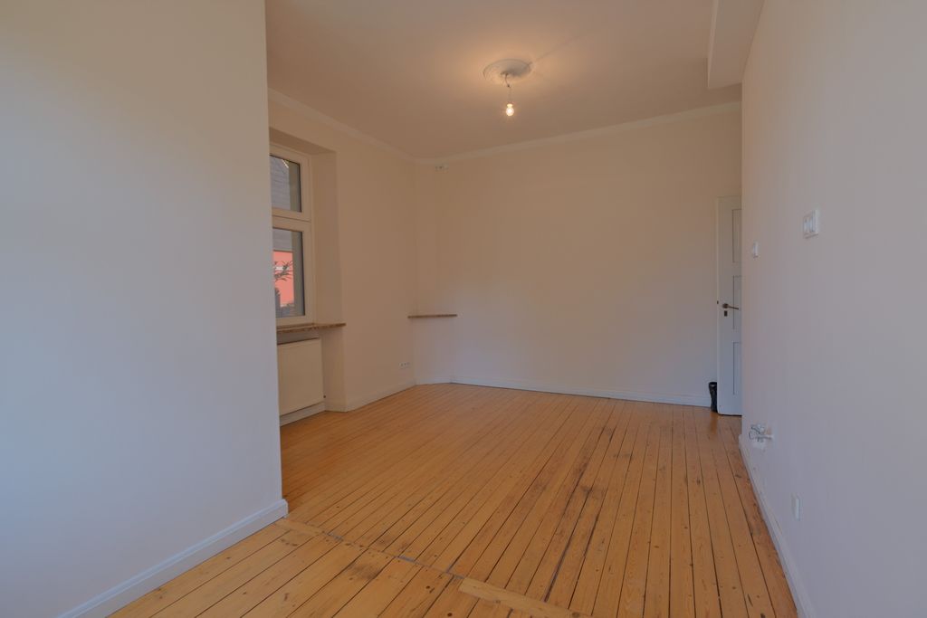 Empty room with white walls, large windows, and a light wooden floor. A bare light bulb hangs from the ceiling.