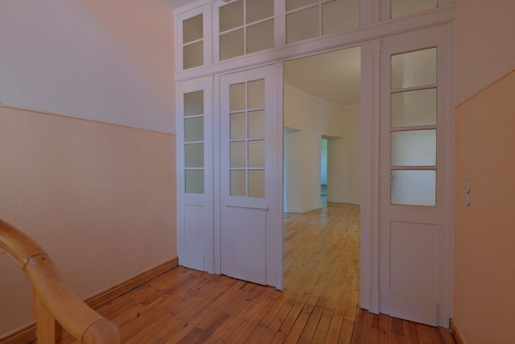 Open white glass doors lead from a hallway with wooden railing into a room with wooden floor and white walls.