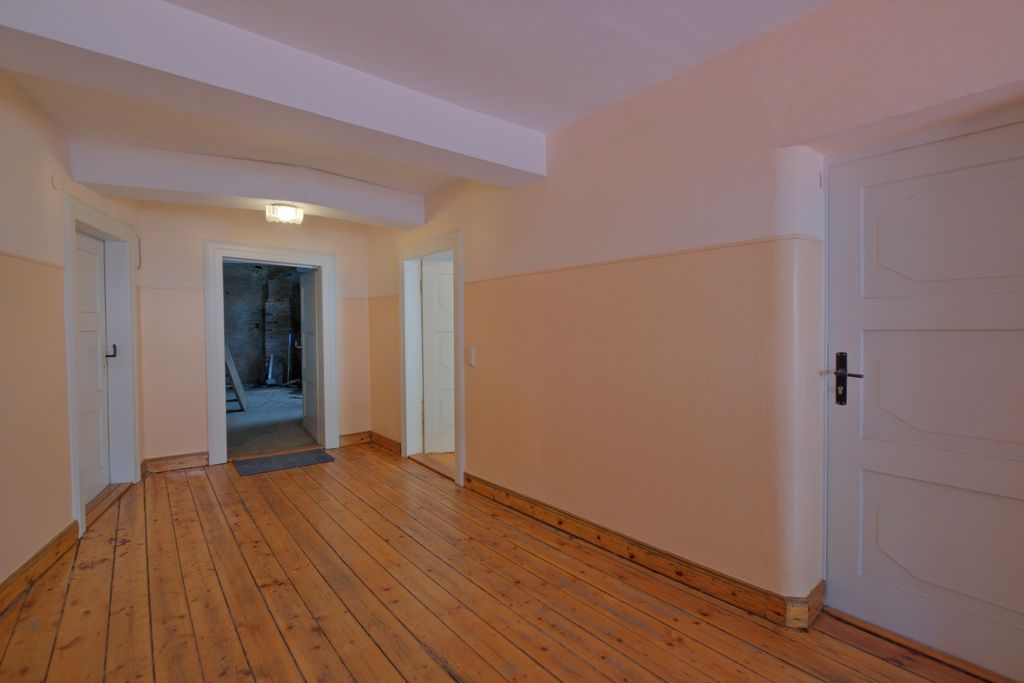 A bright hallway with wooden flooring and several white doors, one leading into a darker room.