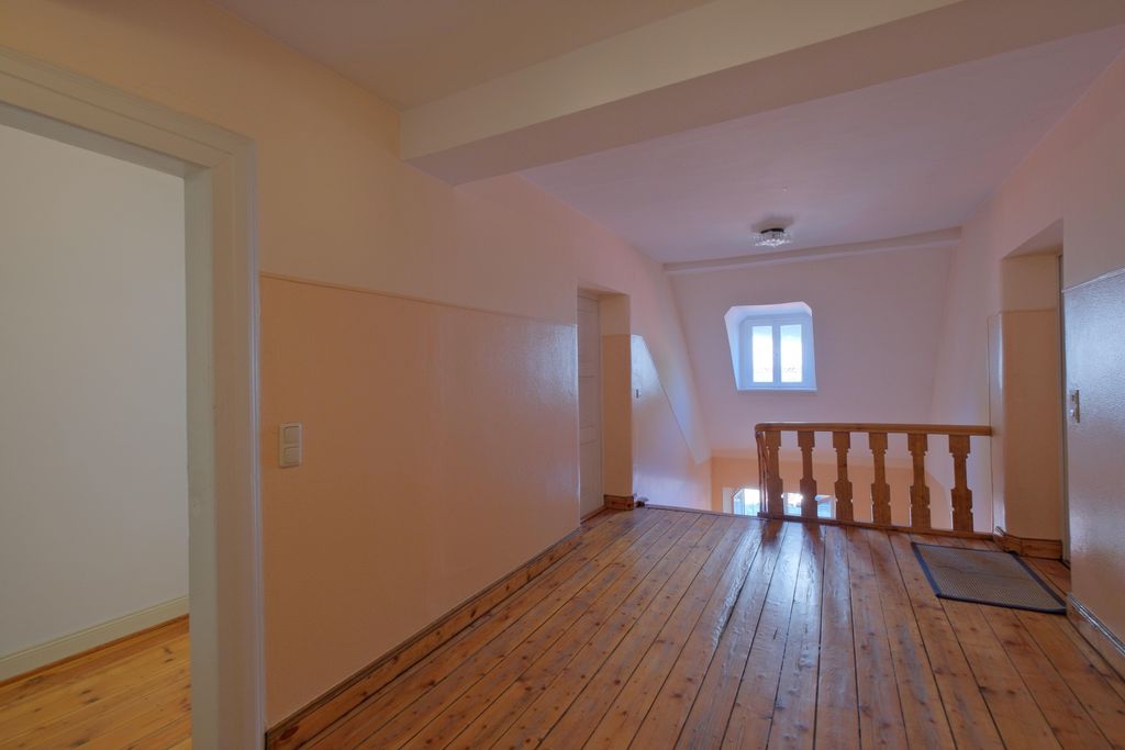 Hallway with wooden floor and light pastel-colored walls in an older house with stairs and a window at the end.