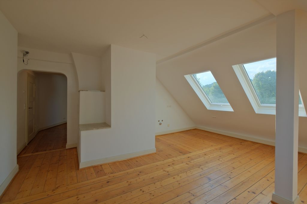 Bright empty attic room with wooden floor, three large skylights, and visible doorway leading to a hallway with white walls.