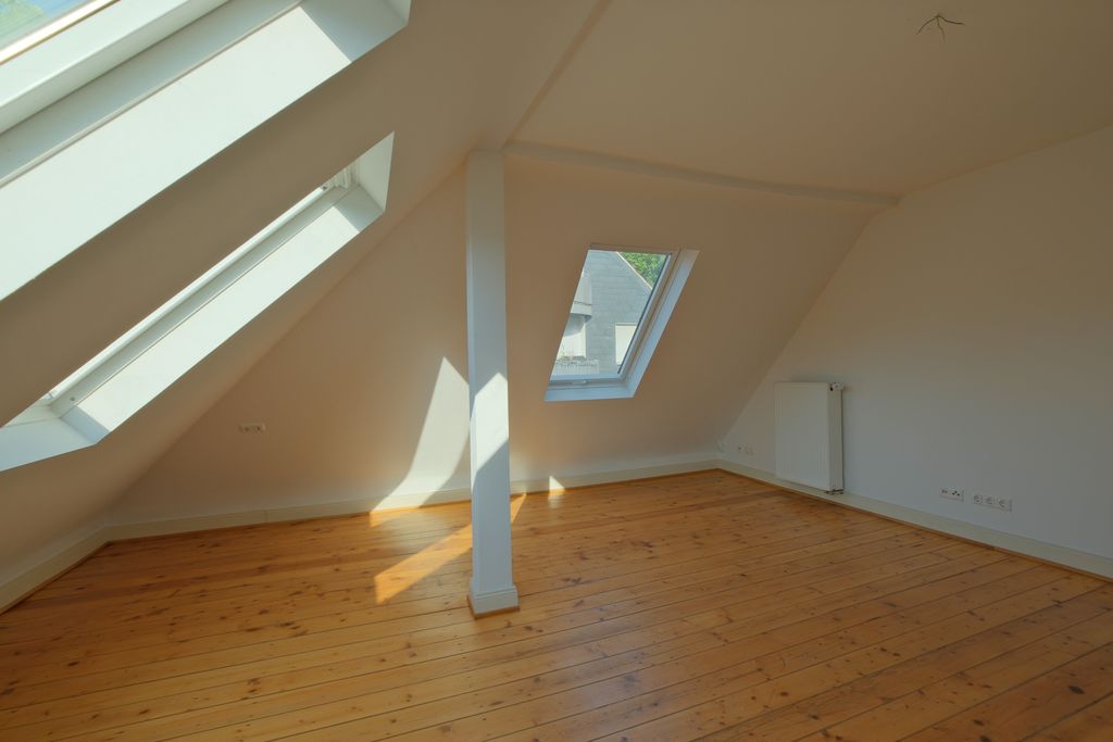 Empty room under a slanted roof with wooden flooring, white-painted walls, a supporting pillar in the center, and several skylights letting in daylight.