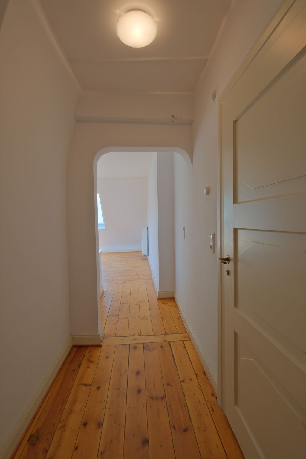 Hallway with wooden floor and white painted walls, leading through a rounded archway into another room with a window and radiator.