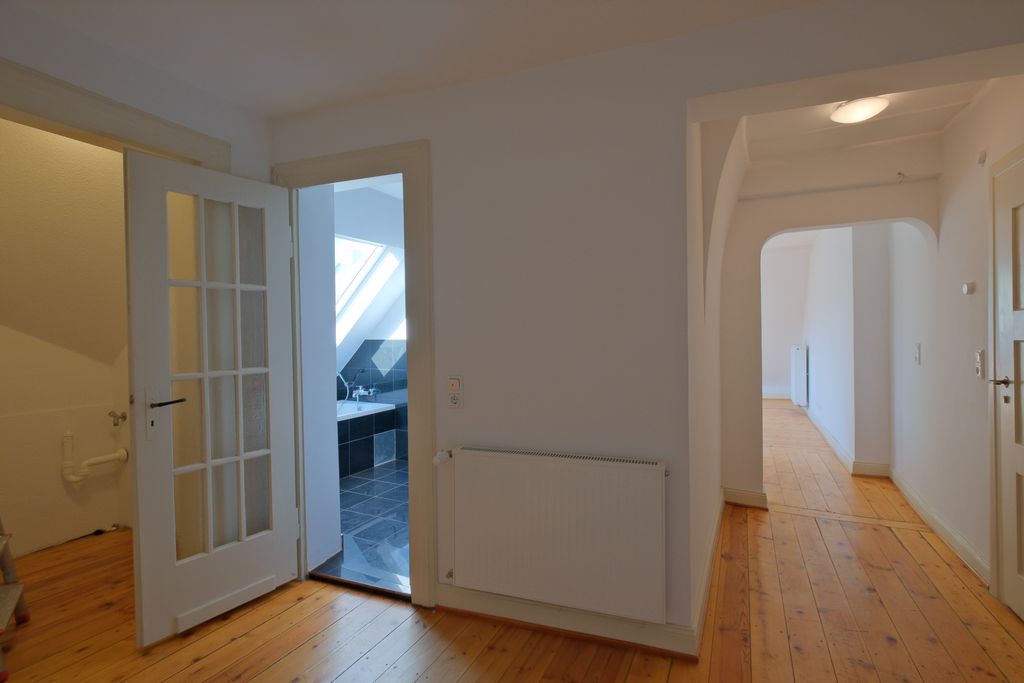 Hallway with light hardwood floor and white walls, open door leading to a bathroom with black tiles and bathtub under a skylight.