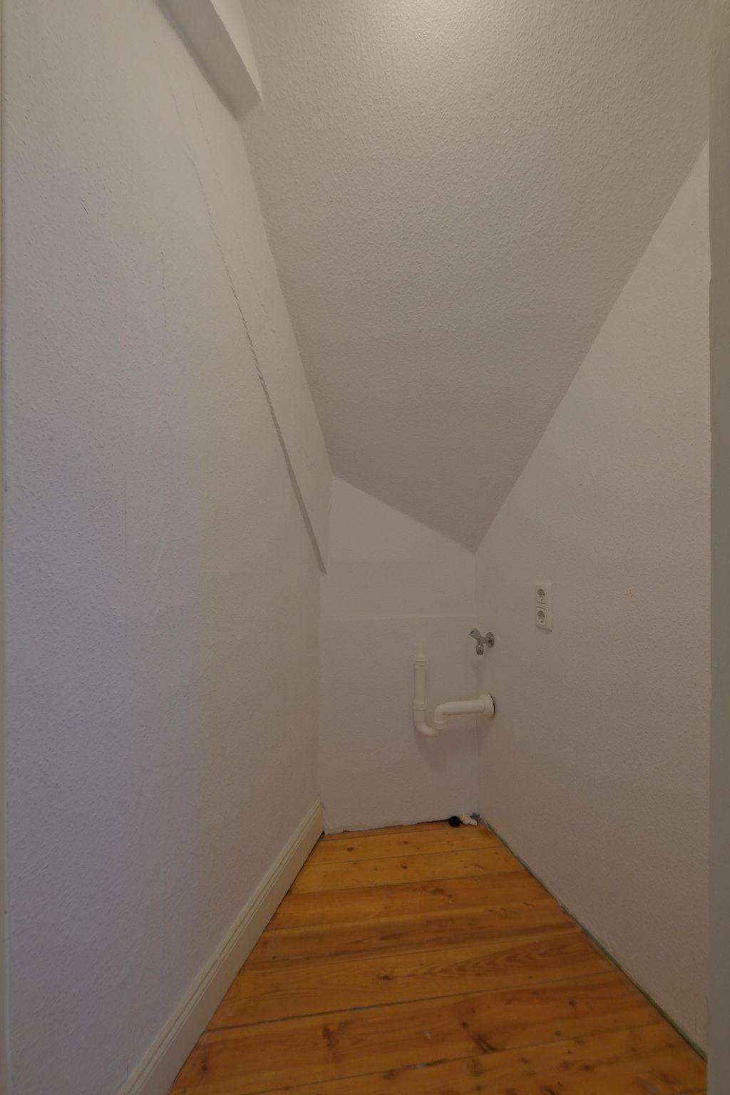 Small empty room with slanted ceiling, white walls, and wooden floor, featuring a water pipe and faucet on the right side.