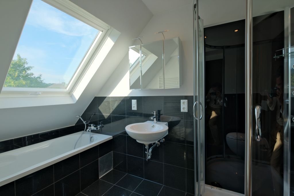 Modern bathroom with black tiled walls, bathtub beneath a large skylight, sink, and glass door shower enclosure.