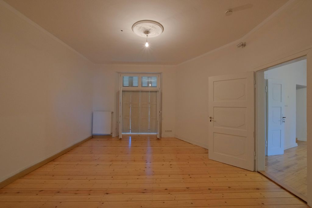 Empty room with wooden floor, white walls, an open white door on the right, and a window with closed blinds at the far end.