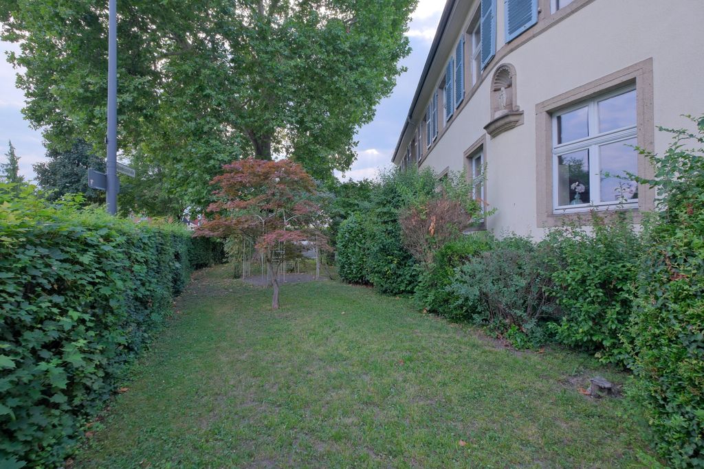 Green garden area beside a light-colored building with blue shutters, surrounded by bushes and trees.