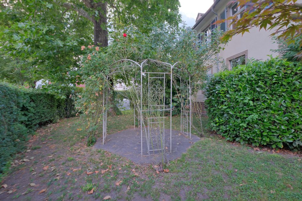 An empty white metal pavilion overgrown with climbing plants in a garden next to a building.