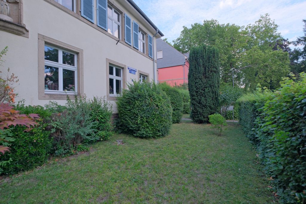 A well-maintained green garden area beside a light-colored building with blue shutters, surrounded by various bushes and trees under a blue sky.