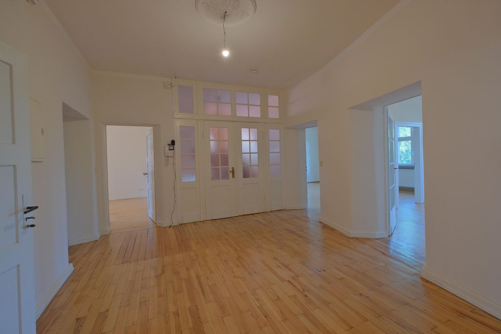Spacious empty room with light wooden floors, white walls, and doors with frosted glass panels leading to adjacent rooms.