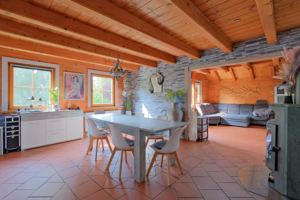 Modern kitchen-living area with wooden beam ceiling, large dining table, white chairs, and an adjoining living room with a gray corner sofa in a space featuring wood and stone walls.