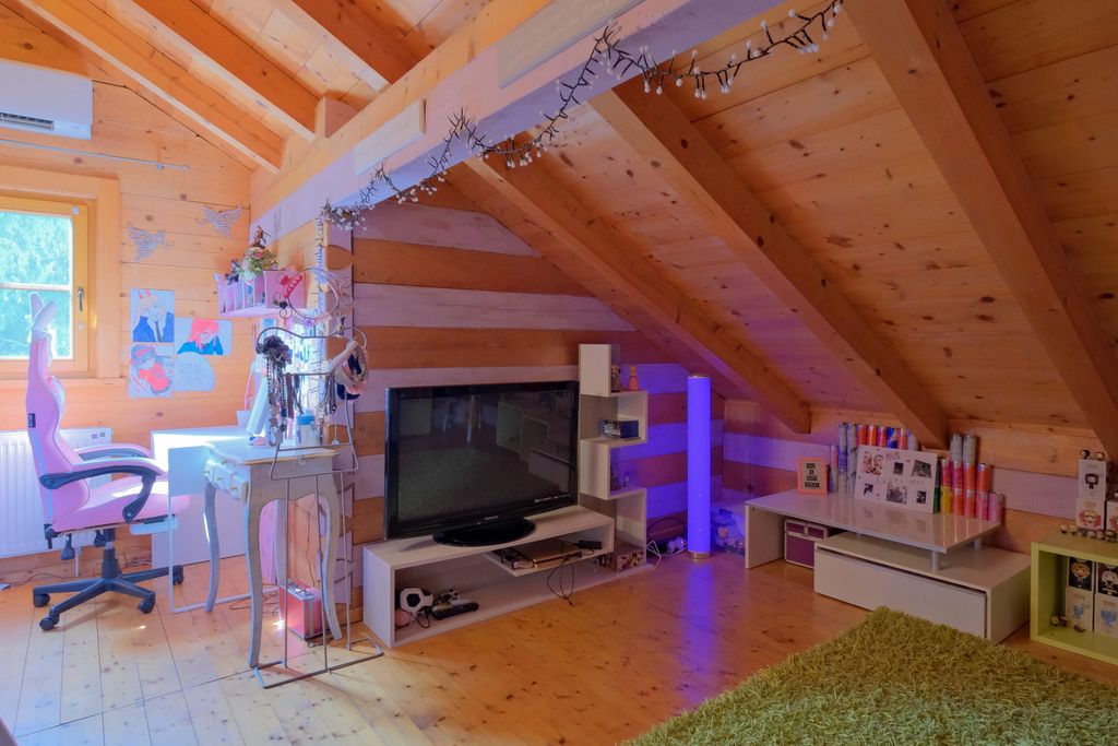 A cozy attic room with wood paneling featuring a pink office chair, a desk with drawings, and a television on a white TV stand.