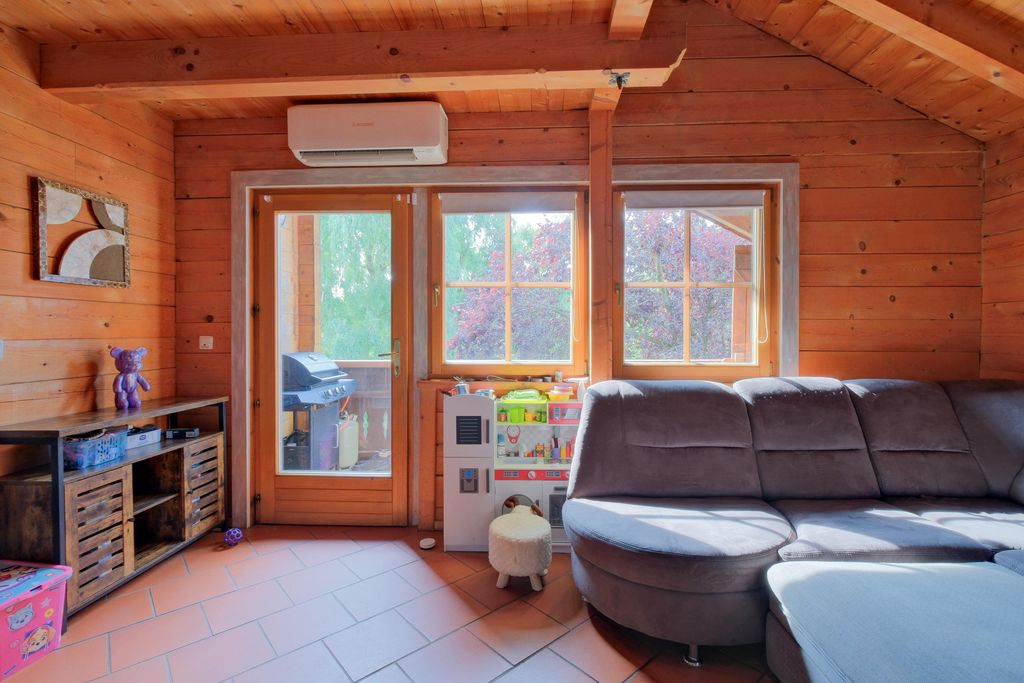 Interior of a cozy wooden cabin with large windows, gray fabric sofa, children's play kitchen, and a terrace with a grill visible outside.