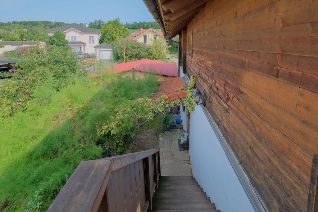 Wooden stairs beside a house with wood paneling, surrounded by green bushes and a view of additional houses in the background.