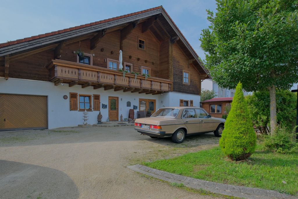 A classic older car parked in front of a traditional two-story house with wooden cladding and a balcony, surrounded by a small garden with trees.