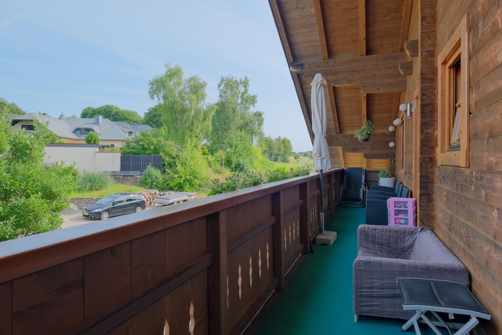 View from a long balcony with wooden floor and railing overlooking green vegetation, houses, and a street with a parked car on a sunny day.