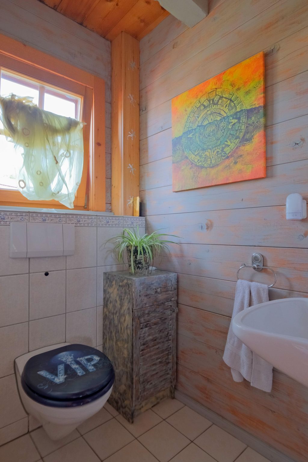 Small bathroom with wooden walls, a wall-mounted toilet featuring a "VIP" toilet seat, a small cabinet with a plant, and a towel next to a sink.