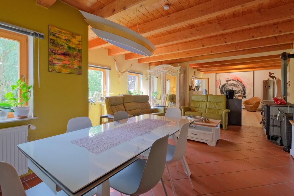 Bright living and dining area with wooden ceiling, white chairs, and a large white dining table against green walls with windows and plants.