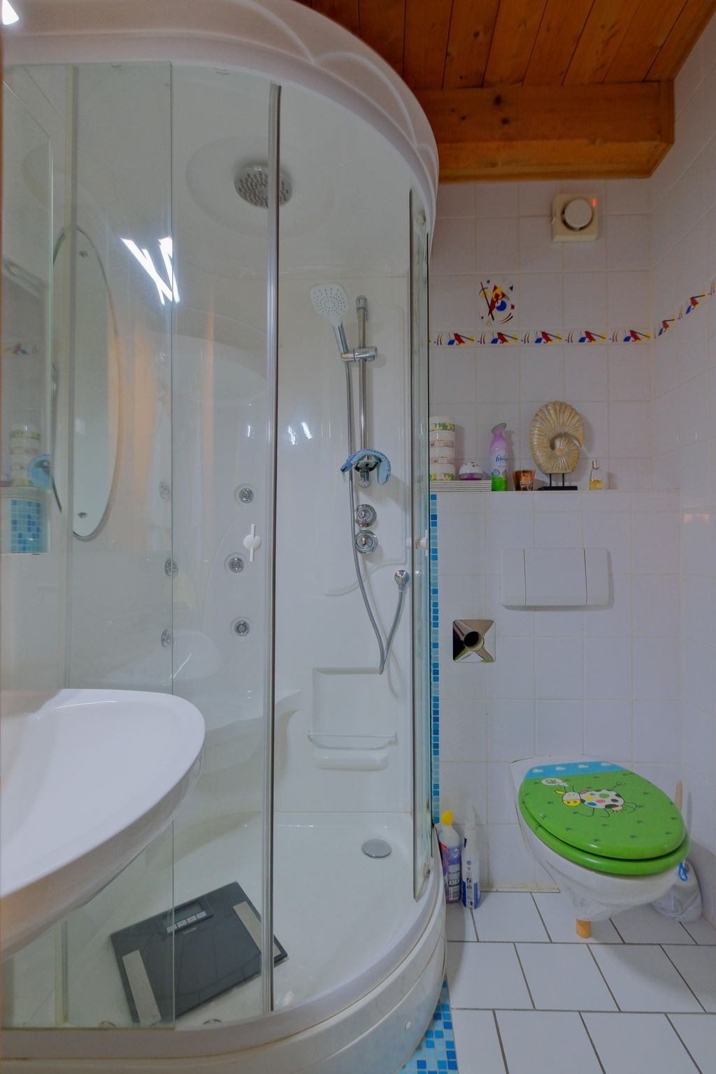 Small bathroom with a rounded shower cabin, a white sink, and a toilet with a green lid featuring a colorful ladybug design, white tiled walls with a colorful border, and a wooden beam ceiling.