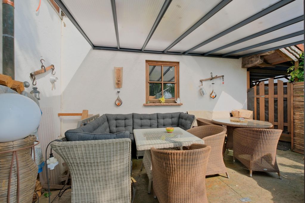 Covered patio featuring a large gray corner sofa, a glass coffee table, several wicker chairs, and a round table against a white wall with a window.