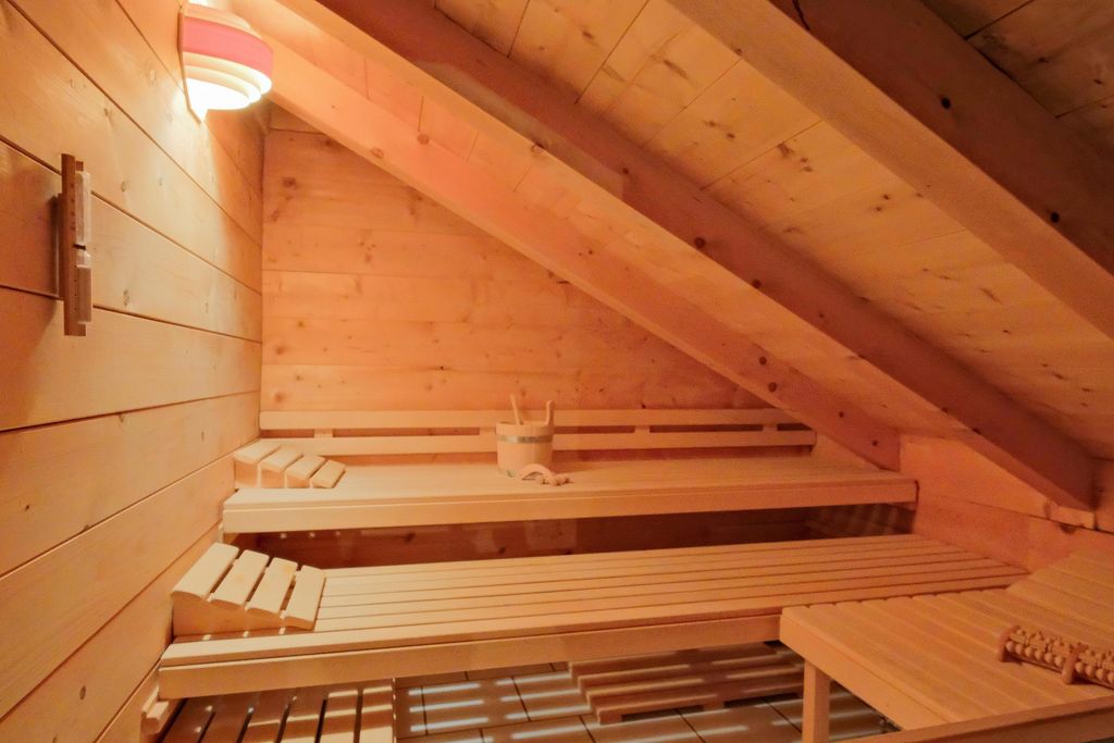 Interior of a small sauna with slanted ceiling and wooden benches, illuminated by a wall lamp.