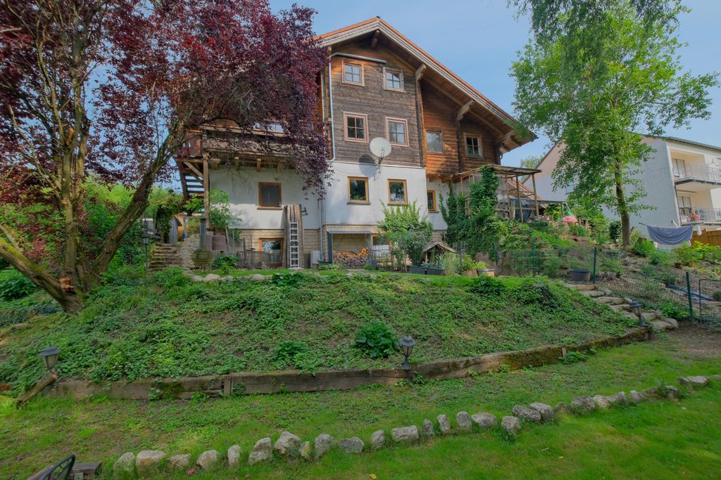 A two-story house with wooden cladding and multiple windows, surrounded by trees and a well-maintained garden with a stone path and plants.