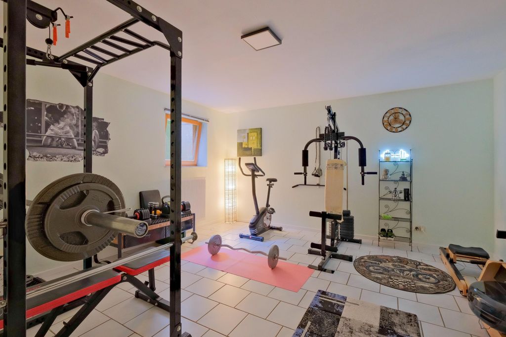 A well-equipped fitness room in a bright space featuring various exercise equipment including a weight rack, exercise bike, weight pulley machine, and a barbell on a yoga mat.
